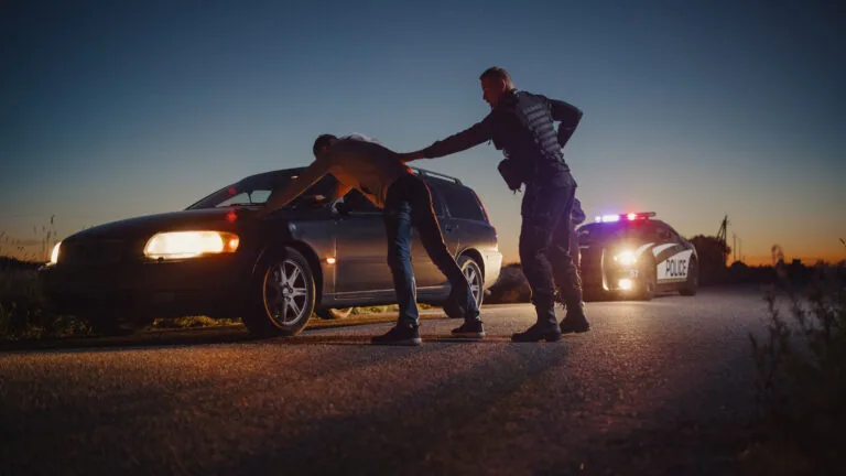 Police officer searches a person by a car at dusk.