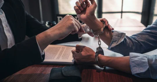Lawyer assisting handcuffed client at a desk.